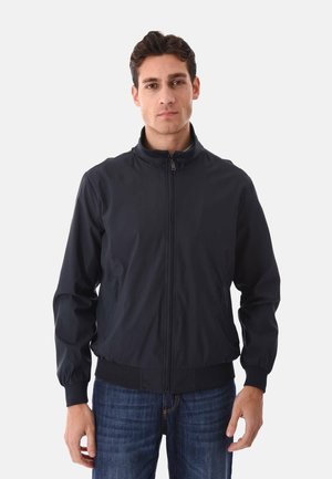 Uomo che indossa una giacca nera con cerniera, polsini e orlo elastici, in piedi su uno sfondo chiaro e uniforme, rivolto verso la fotocamera.