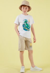 Garçon aux cheveux roux portant un chapeau bob beige, un t-shirt blanc avec un motif de dinosaure, un short beige avec un motif de soleil et des baskets blanches sur fond jaune.