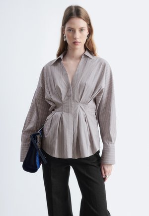 Jeune femme portant un chemisier beige à fines rayures avec manches larges et un pantalon noir, tenant un sac à main bleu marine sur fond blanc.