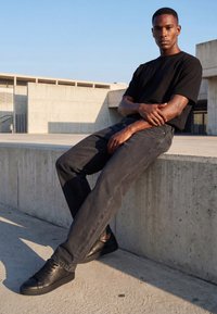 Tânăr purtând tricou negru, blugi negri decolorați și adidași negri, așezat pe o margine din beton într-un mediu urban modern pe timpul zilei.