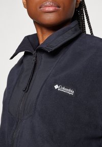 Dunkelblaue Fleecejacke mit Reißverschluss, hohem Kragen und Columbia-Logopatch. Weiche Textur, taillierte Passform und strapazierfähiger Stoff sind deutlich erkennbar.