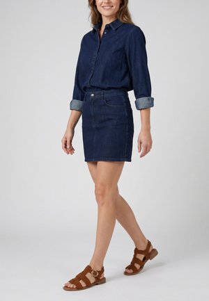 Kvinde går iført en mørkeblå denimskjorte med opkørte ærmer, matchende denimskørt og brune sandaler med stropper og lille hæl.