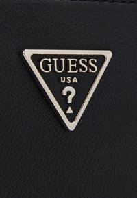 Un materiale testurizzato nero presenta un logo triangolare con accenti dorati, con la scritta "GUESS" e un punto interrogativo al centro.