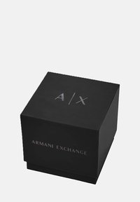 Črna kartonska škatla z besedilom "A/X" na vrhu in "ARMANI EXCHANGE" natisnjeno spredaj v srebrni barvi. Kvadratna oblika, mat zaključek.