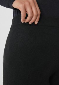 Černé pletené kalhoty s hladkým povrchem a flexibilním pasem, částečně držené rukou. Tkanina vypadá jemně a pohodlně.