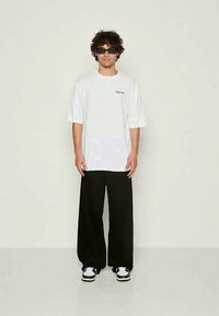 T-shirt blanc oversize avec texte noir ; associé à un pantalon large noir et des sneakers noir et blanc ; modèle portant des lunettes de soleil noires.