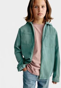 Zelená lehká bunda se dvěma náprsními kapsami, nošená přes růžové tričko, v kombinaci s modrými džínami, na dítěti se středně dlouhými vlasy.