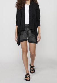 Svart cardigan med subtila glitter, öppen front och långa ärmar, i kombination med mörka denimshorts och svarta sandaler med dekorativa spännen.