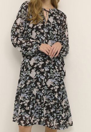 Kvinde iført en sort kjole med lange ærmer med et let blomsterprint og bindebånd i halsudskæringen, stående med hænderne blidt foldet.
