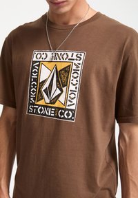 Brun bomulds t-shirt med et stort grafisk print i sort og gul med en diamantform og teksten "Volcom Stone Co." i fed skrift.