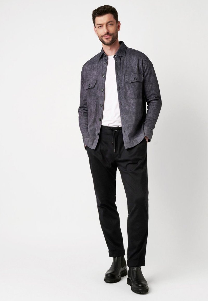 Donkergrijze button-up shirt voor heren met twee borstzakken, gelaagd over een wit T-shirt, gecombineerd met zwarte broek en zwarte enkellaarzen.