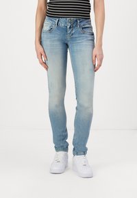 Jeans ajustados en denim azul claro con un acabado desgastado, que presentan un diseño de cinco bolsillos y doble botón en la bragueta, combinados con zapatillas blancas.