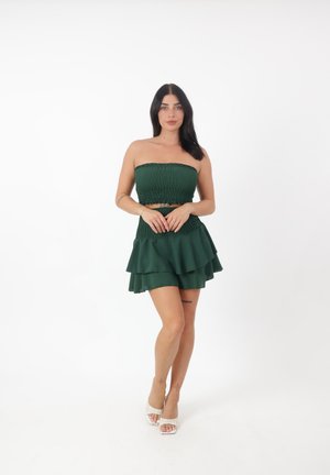 Grünes, gestuftes Kleid mit gerafftem Oberteil, schulterfreiem Design und Rüsche am Saum. Der Stoff hat eine glatte Textur und wird mit hellen Absätzen getragen.