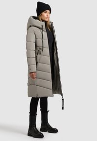 Abrigo largo tipo puffer en gris claro con capucha, diseño acolchado, bolsillos laterales y cremallera. Combinado con leggins negros y botas tipo botín.