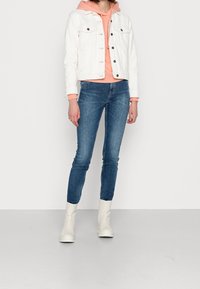 Biała dżinsowa kurtka na różowej bluzie z kapturem, niebieskie obcisłe dżinsy i białe botki za kostkę. Stylizacja wyróżnia się kontrastującymi kolorami i fakturami.