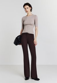 Top a coste beige con maniche corte, abbinato a pantaloni a campana marrone scuro. La modella tiene una borsa nera, design minimalista, texture liscia.