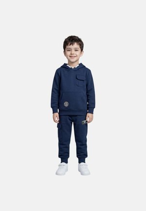 Garçon souriant debout, portant un sweat à capuche bleu marine avec des poches et un pantalon de jogger assorti avec des patchs, ainsi que des baskets blanches sur fond blanc.