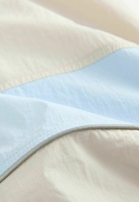 Teksturowana tkanina w delikatnych odcieniach beżu i jasnego niebieskiego, z gładkim wykończeniem i łagodnym wygięciem, podkreślająca wykończone szwy.