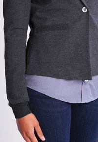 Dark gray, fitted blazer in soft material with a single button closure and pocket detail, layered over a light blue shirt.