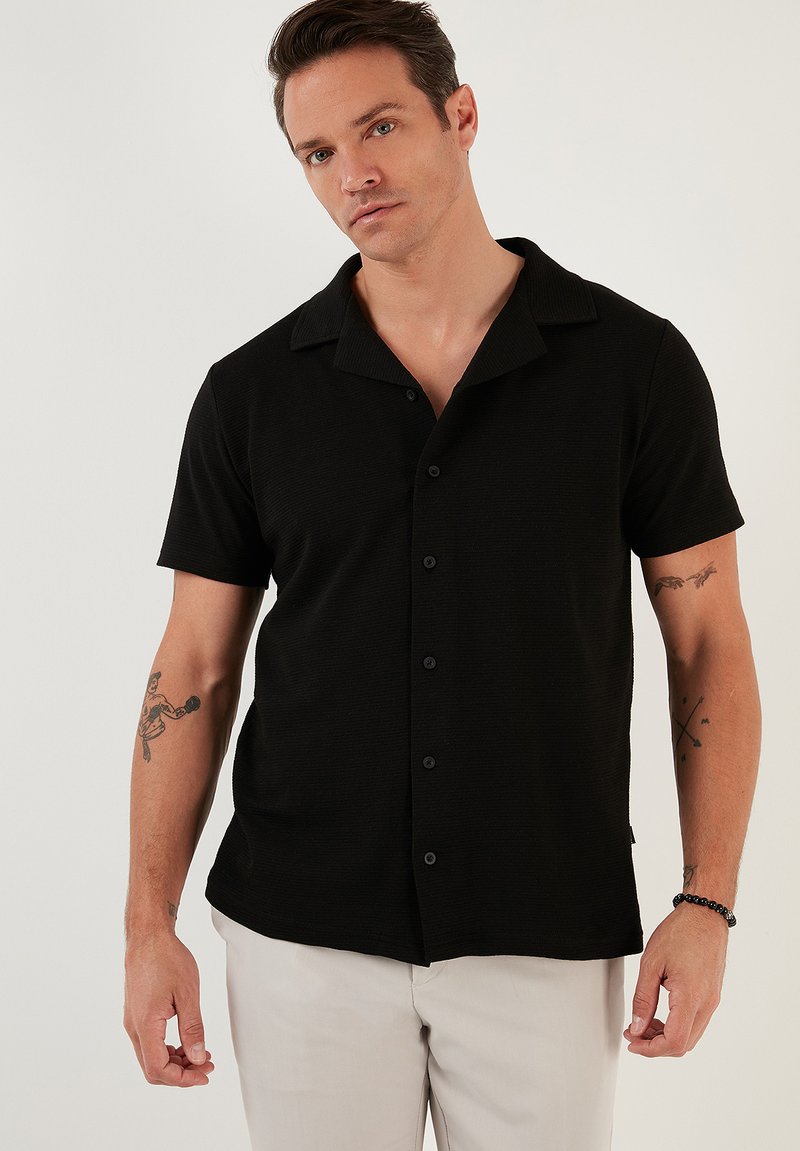 Camisa negra de manga corta con una tela texturada, botones al frente y un cuello clásico, combinada con pantalones de color claro.