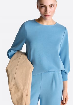 Mladá žena oblečená v svetlomodrej blúzke s dlhými rukávmi a nohaviciach, drží béžovú bundu cez ľavé rameno na bielom pozadí.