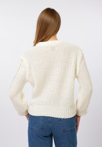Hvid strikket sweater med løs pasform, der har tekstureret syning, ribbet kant og faldende ærmer. Kombineret med blå højtaljede jeans.