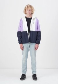 Kleurgeblokte windbreaker in wit, lavendel en marineblauw. Voorzien van een ritssluiting aan de voorkant, elastische manchetten en een logo op de linkerborst. G gedragen over een zwart shirt.