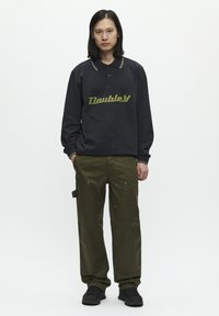 Schwarzer Pullover mit einem grünen "Doublet"-Logo, kombiniert mit olivgrünen Cargo-Hosen mit Taschen und sichtbaren Nähten. Schwarze Sneaker runden den Look ab.