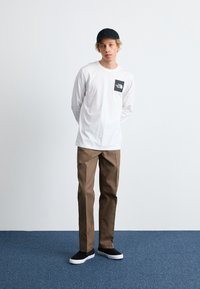 Weißes Langarmshirt mit schwarzem Logopatch, braune Hose und schwarze Sneakers mit weißen Sohlen, vor einem neutralen Hintergrund.
