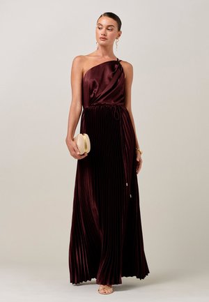 Kobieta w ciemnoczerwonej, plisowanej sukni bez rękawów, trzymająca małą beżową kopertówkę, nosząca złote kolczyki i sandały, na jednolitym tle.