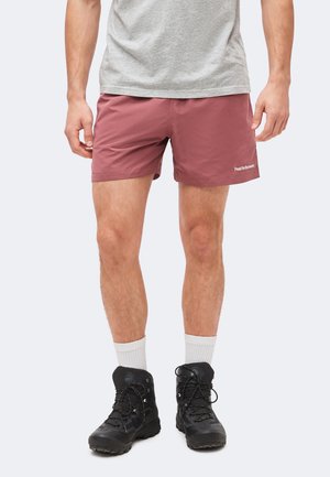 Άνδρας που φοράει σκόνινα ροζ σορτς, γκρι μπλουζάκι, λευκές κάλτσες μέχρι τον αστράγαλο και μαύρα ορειβατικά μποτάκια, στέκεται μπροστά σε ανοιχτό φόντο.