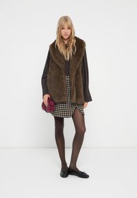 Vrouw in bruine bontvest, zwarte blouse, geruite mini-jurk, zwarte panty's, zwarte ballerina's, met een kleine bordeauxrode handtas, staand op een witte achtergrond.