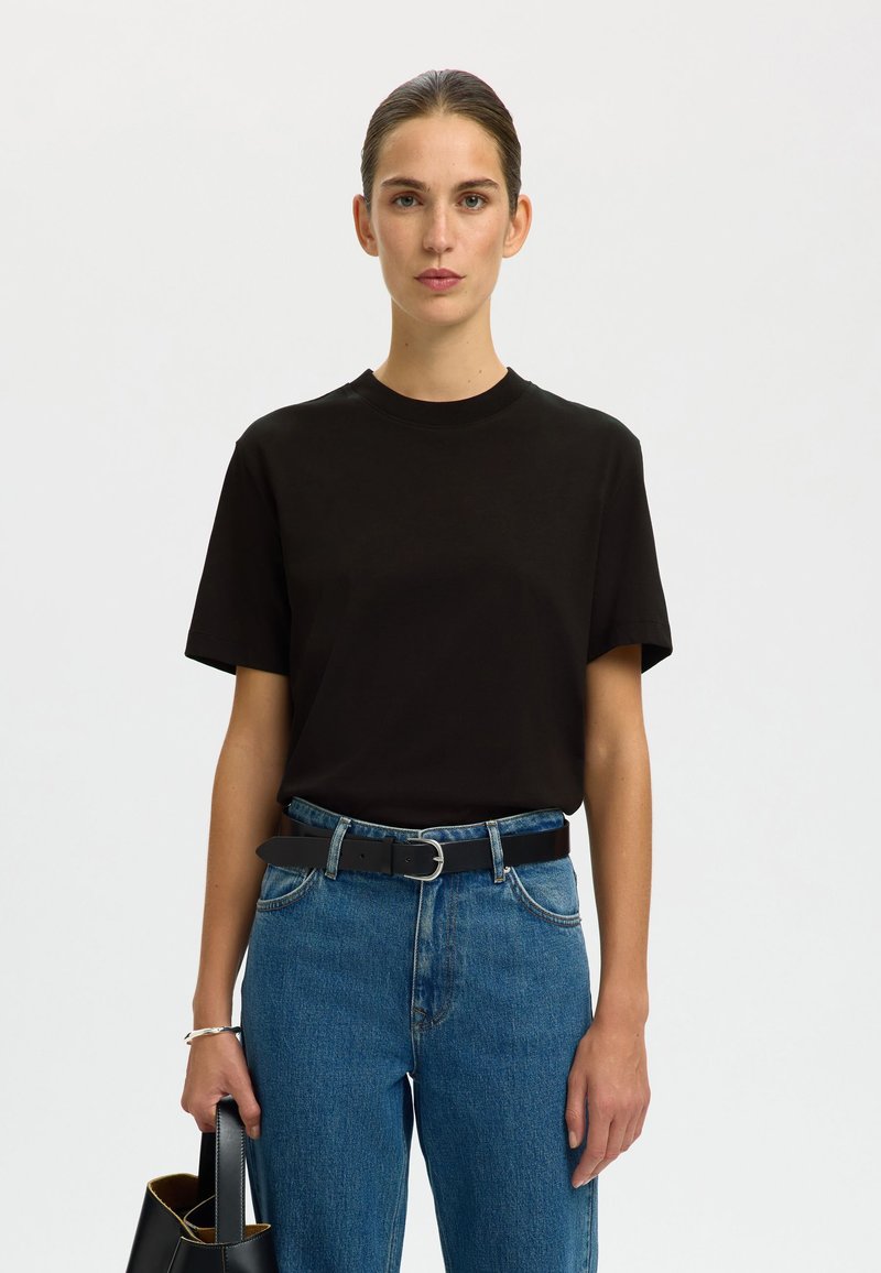 Juoda medvilninė t-shirt su trumpomis rankovėmis, apvaliu kaklu, derinama su aukštai pjaustytomis mėlynomis džinsomis ir juoda diržu. Modelis laiko juodą rankinę.