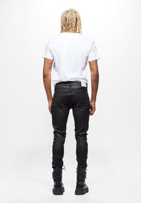 Persona con dreadlocks rubios, vistiendo una camiseta blanca, pantalones ajustados negros y botas negras con hebilla, de pie de espaldas sobre un fondo blanco.