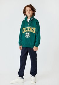 Zielona bluza z połową zamka, logo "CHALLENGER" w kolorze żółtym, zestawiona z granatowymi spodniami dresowymi i białymi sneakersami. Gładki materiał, luźny krój.