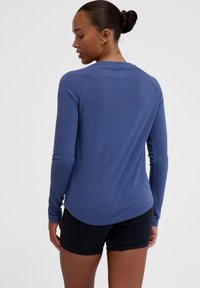 Długo rękawowa koszulka sportowa w kolorze granatowym, wykonana z miękkiego materiału, z okrągłym dekoltem, raglanowymi rękawami i lekko zaokrąglonym dołem. Widoczne czarne szorty.