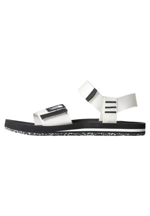 Sandalias blancas y negras con correas ajustables, una suela texturizada negra y un borde blanco moteado. Presenta un logo en la correa.