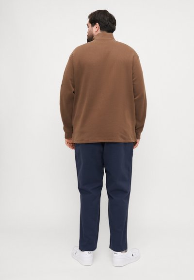 Bruine sweatshirt met trechterhals en lange mouwen, gedragen over een marineblauwe broek en witte sneakers, gezien vanuit de achterkant tegen een effen achtergrond.