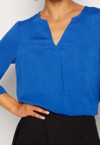 Niebieska, teksturowana bluzka z dekoltem w serek i rękawami 3/4, połączona z czarnym pasem. Skoncentruj się na gładkim wykończeniu materiału.