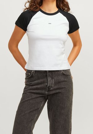 Nainen, joka pukeutunut istuvaan valkoiseen t-paitaan mustilla raglanhihnoilla ja mustalla "JJXX"-logolla, yhdistettynä korkeavyötäröisiin haalistuneen mustiin farkkuihin.