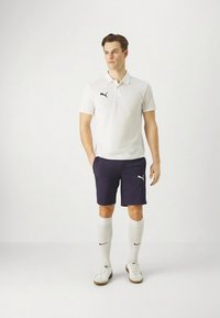 Hvit polo med svart logo, marineblå shorts, hvite knestrømper og hvite joggesko med brune såler. Enkel, atletisk design.