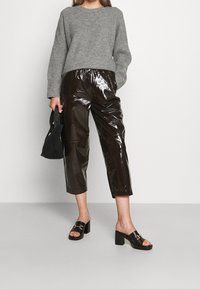 Femme portant un pull en maille gris, un pantalon court en vinyle noir brillant, des mules noires à talons ouvertes et tenant un petit sac à main noir.