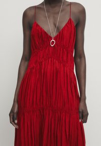 Punainen hihaton mekko, jossa on syvä V-kaula-aukko, kerrostettu tekstuuri ja elastinen vyötärö. Ominaisuuksiin kuuluvat ohuet olkaimet ja liehuva, kerrostettu helma. Hopeinen riipuskaulaketju.