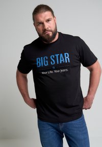 Camiseta de algodón negra con texto azul "BIG STAR" y la frase "Tu vida. Tus jeans." El modelo lleva unos jeans azules, con un fondo sencillo.