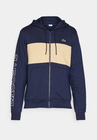 Felpa con zip color navy, con una striscia orizzontale crema, dotata di cappuccio con cordino, tasche frontali e logo sulla manica. Materiale in misto cotone.