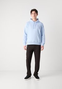 Light blue hoodie with a front pocket, adjustable drawstring, and ribbed cuffs, paired with black trousers and dark sneakers.