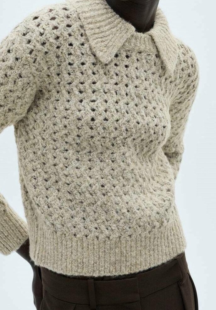 Beżowy sweter z dzianiny o teksturowanym wzorze koronkowym, z prążkowanym dołem i kołnierzem. Wykonany z miękkiej przędzy, z niewielkimi różnicami w kolorze.