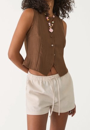 Femme portant un haut marron sans manches boutonné, un short beige avec cordon et un collier multicolore en perles avec un pendentif rose.