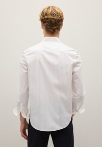 Camicia bianca con righe verticali sottili, maniche lunghe arrotolate ai polsi, orlo curvo e dettagli con bottoni sui polsi.