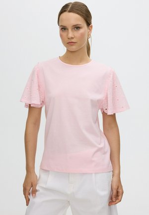 Joven con cabello castaño claro recogido en una coleta, que lleva una camiseta rosa claro con mangas cortas de encaje y pantalones blancos sobre un fondo liso.